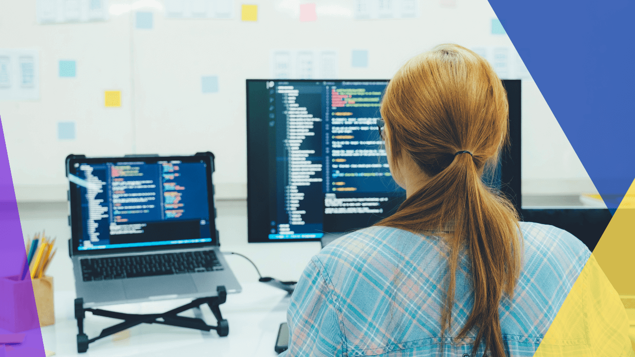 A woman in a blue plaid shirt with her hair in a ponytail sits at her desk with two computers displaying lines of code, one on a stand and one on the desk.