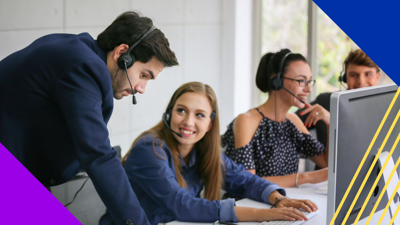 A man wearing a headset leans over to speak with a woman, while two others with headsets work at a computer in the background.