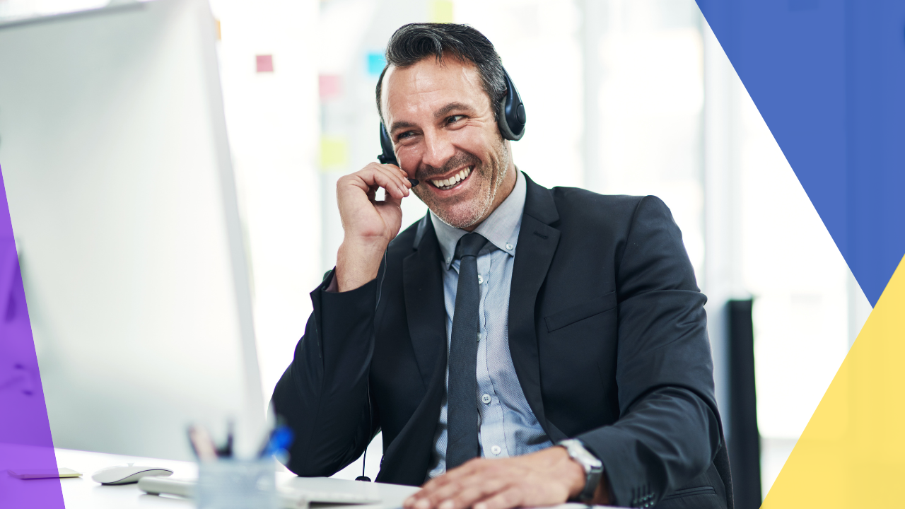 A man wearing a suit and headset smiles while working at his desk.