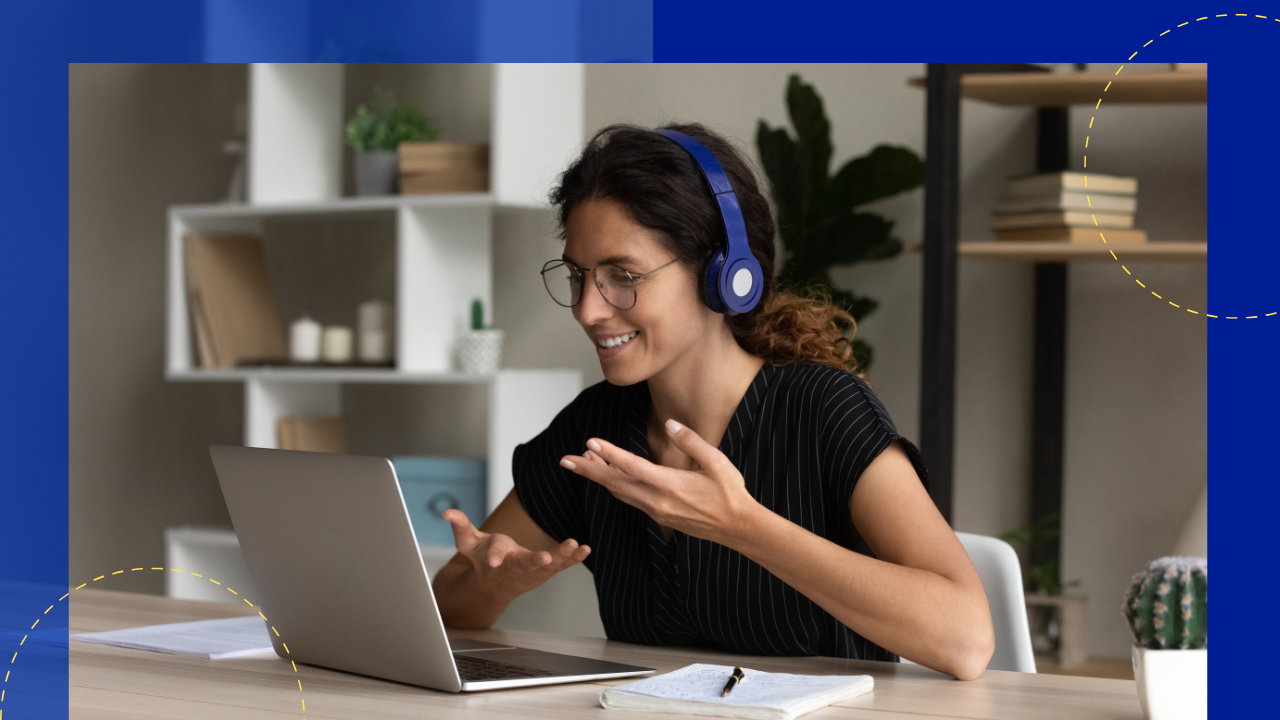 A woman wearing headphones smiles and gestures while sitting in front of an open laptop. She is in a home office setting with a shelf and a plant in the background.