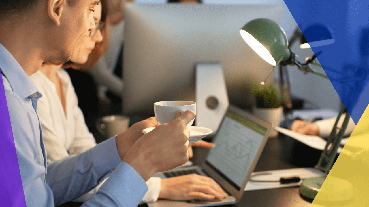 A person in a blue shirt drinks coffee while looking at a laptop with a colleague, surrounded by a desk lamp, papers, and a plant.