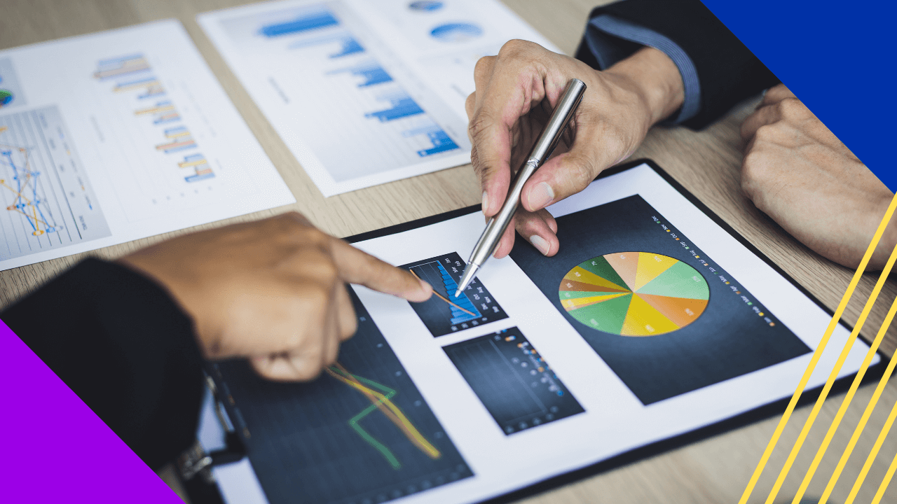 Two people are reviewing data on a page of charts and graphs, with one pointing to a chart using their finger and the other with a silver pen.