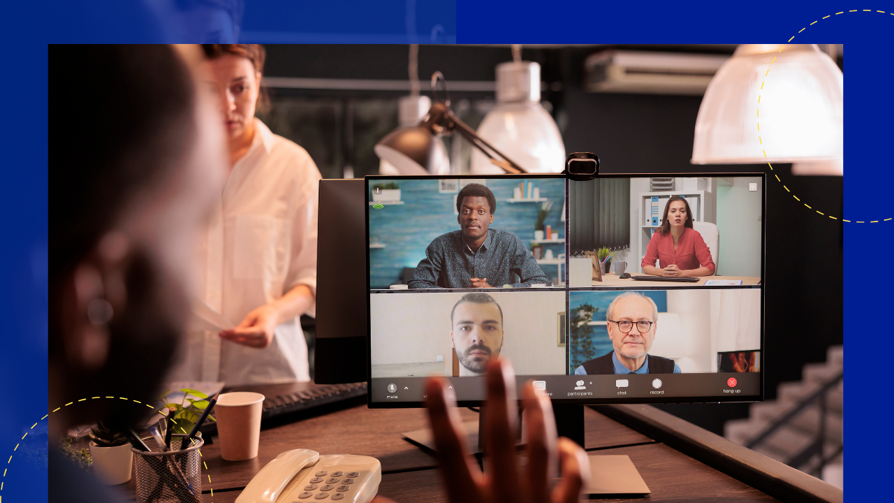 A group of people are gathered around a table in a dimly lit room, attentively watching a video call on a large screen while a presentation is displayed on a laptop in front of them.