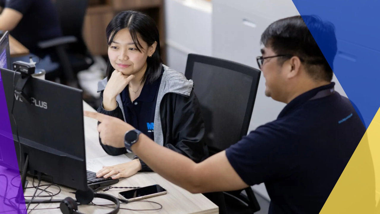 A man and a woman are seated at a desk in front of a computer. The woman is looking at the screen while the man is looking at the woman. The man has a hand on the woman's arm, as if he is pointing something out to her. The man is wearing a black shirt and the woman is wearing a gray and blue jacket. The woman has her hand on her chin and the man has his other arm outstretched. The man is wearing a watch on his wrist. There is a phone and a headset on the desk in front of them.