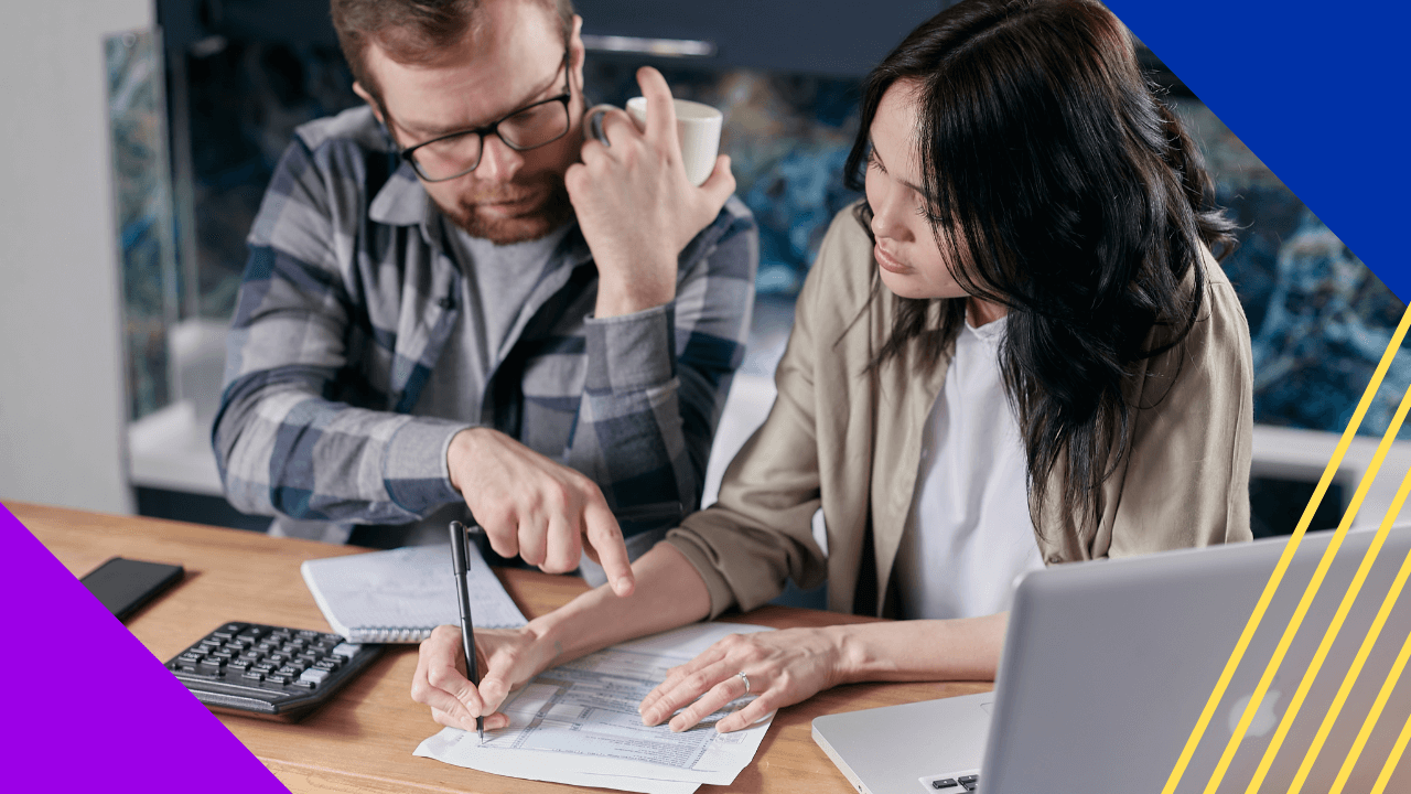 A woman with long dark hair and a man with a beard and glasses sit at a table, casually dressed, as the woman fills out a document with a pen.