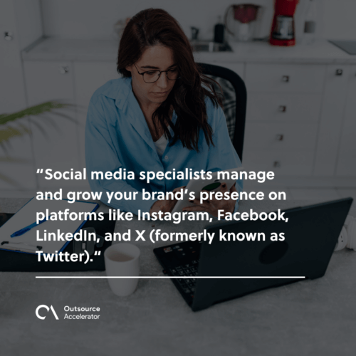 A woman in a blue shirt is sitting at a table with a laptop, coffee cup, and phone, working on her social media.