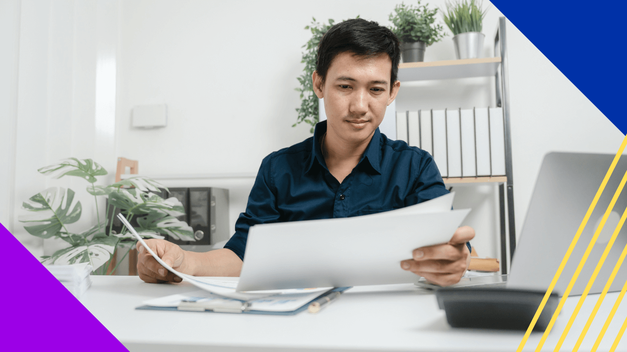 A man in a blue shirt sits at a desk, focused on reviewing documents, with a laptop, paperwork, plants, and a bookshelf in the background.