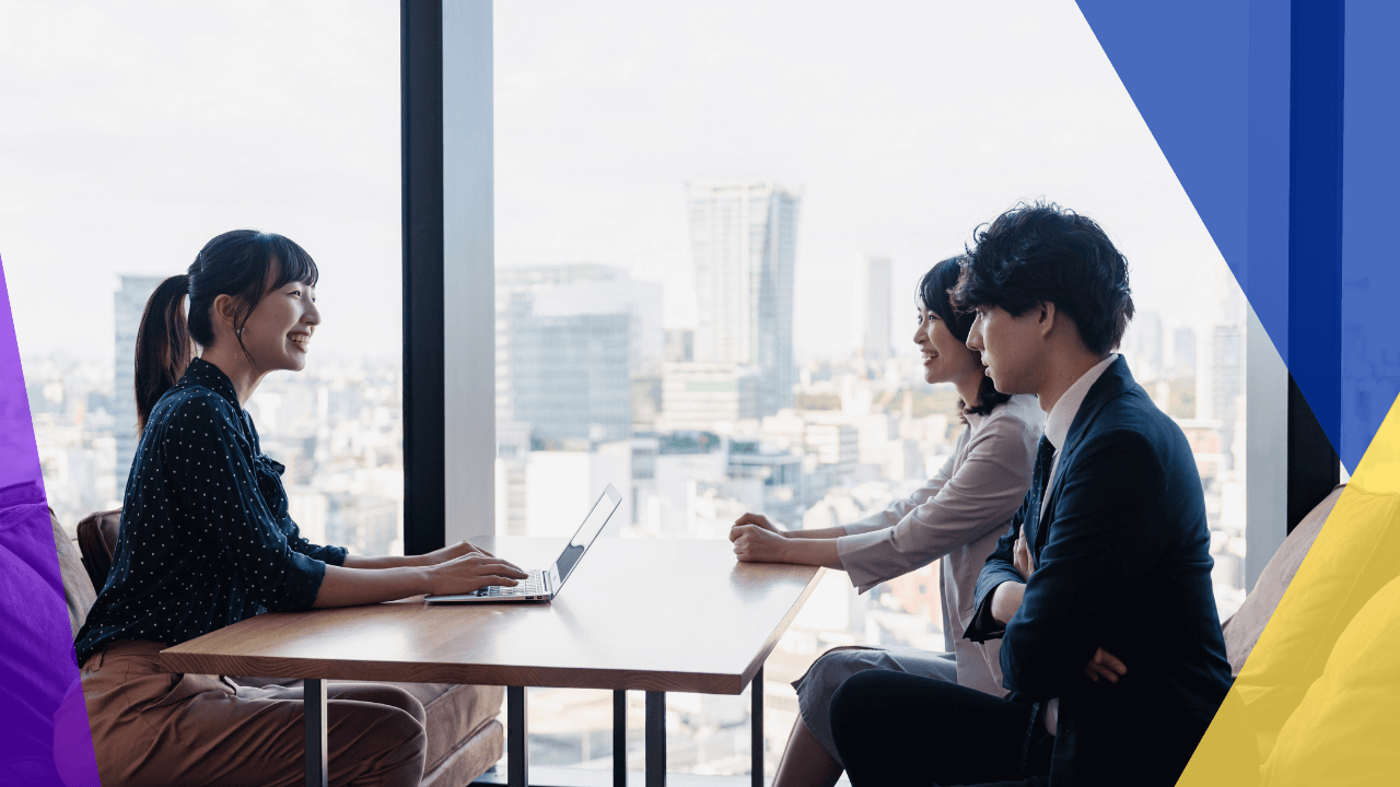 Three young professionals sit at a table in a modern office with a city skyline in the background. A woman in a polka-dot blouse smiles while using a laptop, while a woman in a white blouse and a man in a suit engage in conversation.