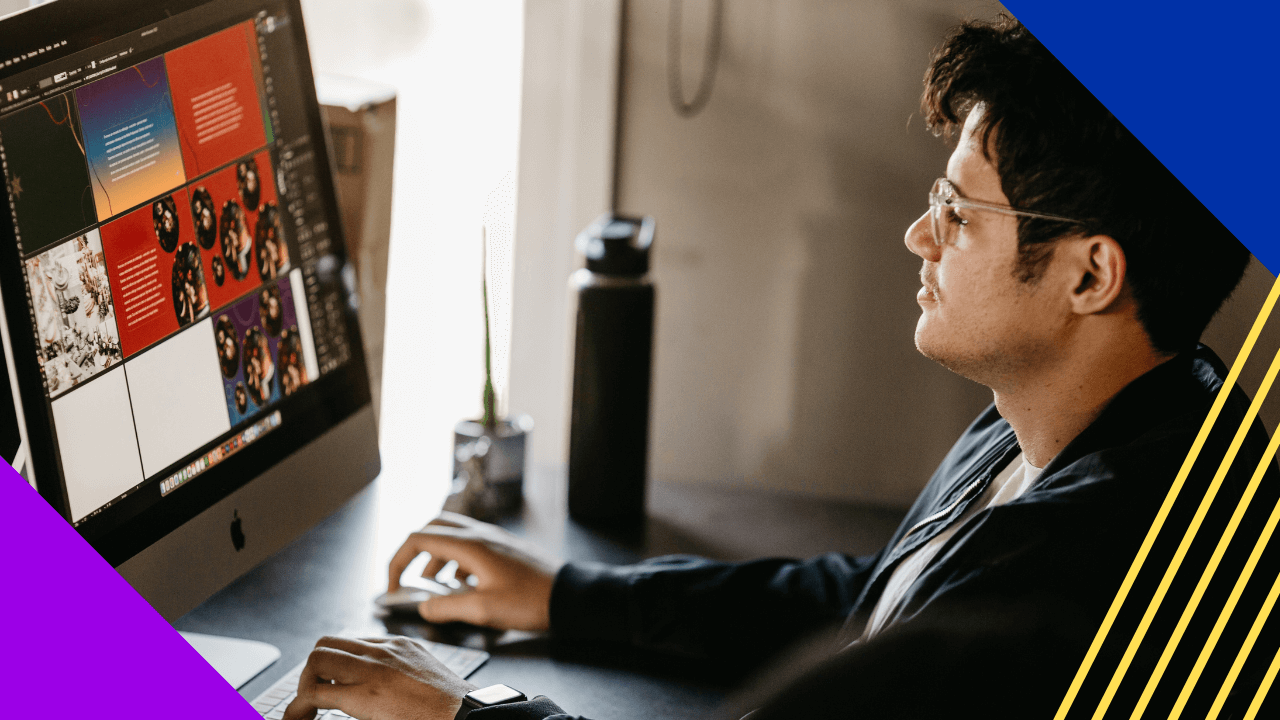 A young man wearing glasses works intently at a computer desk, viewing a vibrant digital design on a large monitor. His well-lit workspace includes a small green plant and a water bottle.